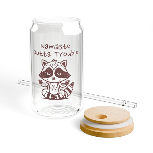 Clear glass tumbler with a raccoon illustration and 'Namaste Outta Trouble' text, accompanied by wooden lid and a straw on a white background.